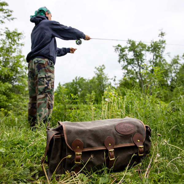 A man fishing along a river bank with a soft sided tackle box sitting next to him. 