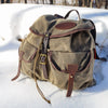 An waxed canvas backpack with dark brown leather trim and hardware, resting on a bed of white snow outdoors.