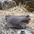 A small canvas pouch with a zipper and a round embossed leather logo patch sits on a log outdoors near dry grass and water.