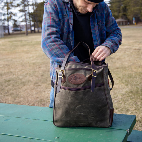 A man is placing items into a dark brown waxed canvas tote bag with brown leather trim, standing on a green wooden picnic table outdoors.