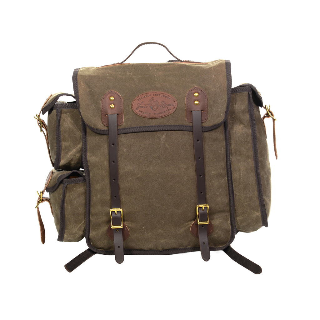 A waxed canvas backpack with dark brown leather straps and trim, featuring a central flap pocket with brass-tone buckles and a circular leather logo patch.