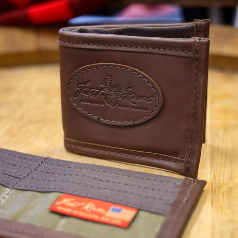 A brown leather bifold wallet with an embossed oval logo on the front, shown standing upright on a wooden surface. A second wallet is partially visible in the foreground, showing a gray fabric interior and a small red tag.