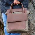A person outdoors holding a brown leather and canvas tote bag. The bag has a large brown leather front flap with a debossed logo, a front pocket, top handles, and side gold-tone hardware.