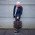 A man holding a waxed canvas and leather briefcase as he walks own the sidewalk. 
